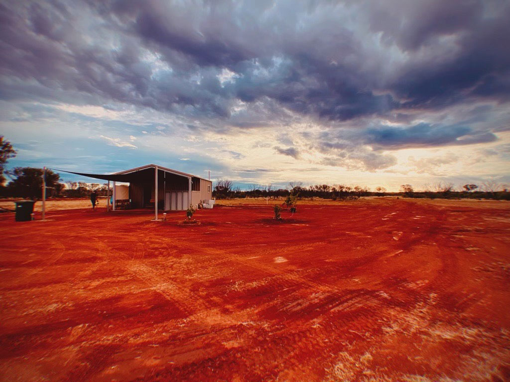 Aboriginal housing in Aboriginal hands -- image shows a small house with a shadecloth verandah in a desert landscape with moody clouds above