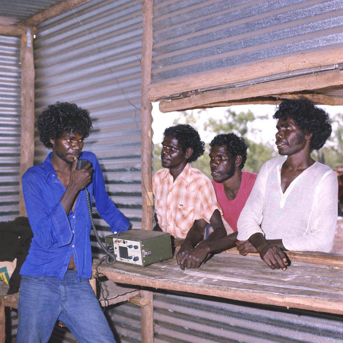 A young Aboriginal man stands talking into a radio in a bush-pole and corrugated iron structure with an open window. Standing outside at the window are three more young Aboriginal men, looking at the man inside.