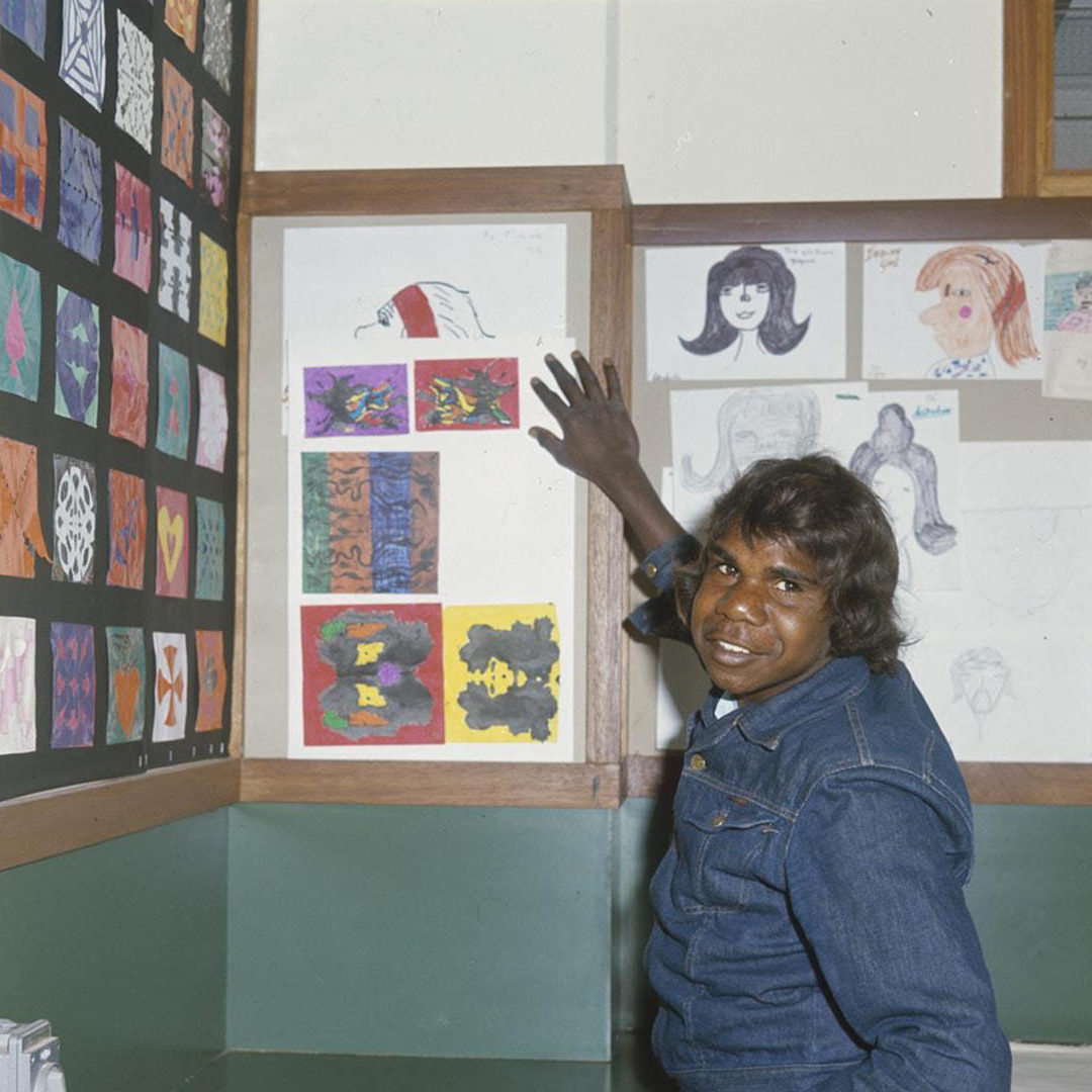 Young Aboriginal man in a denim jacket with his arm extended behind toward children's artwork on the wall of a school