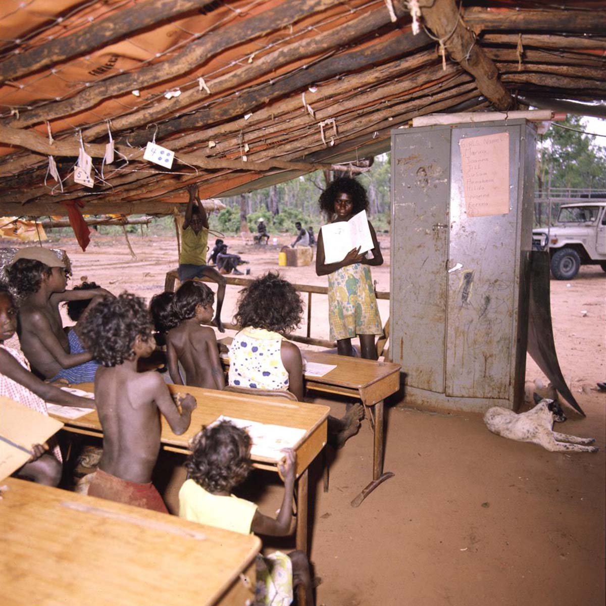 Under a hand-built open-sided timber shelter, a young Aboriginal woman stands showing an open book to a class of at least eight young students who are sitting and standing behind small desks. Next to the teacher is a tall metal cabinet. A dog lies in front of the cabinet and in the background is a land rover and several seated adults.