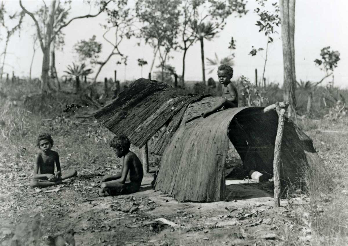 Children are seated on the ground next to and (in one case) atop a simple bark shelter